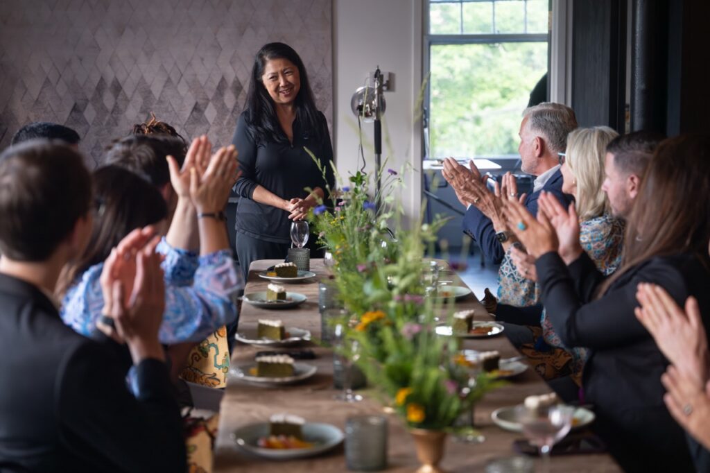Woman addresses seated guests at a long table with floral centerpiece.