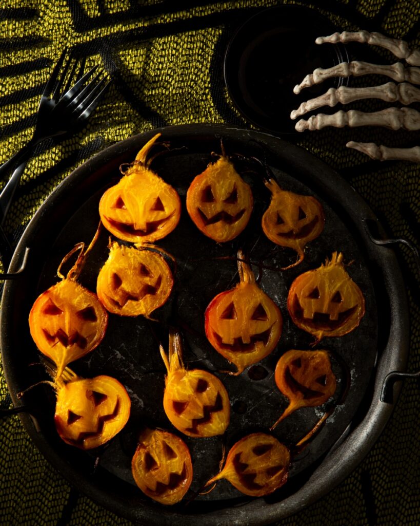 A tray of roasted golden beet halves carved with jack o’lantern faces and drizzled with apple cider glaze on a platter.