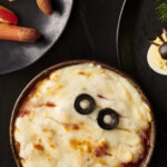 A round bowl of pizza dip with strips of white cheese over top and two olives to look like a mummy.