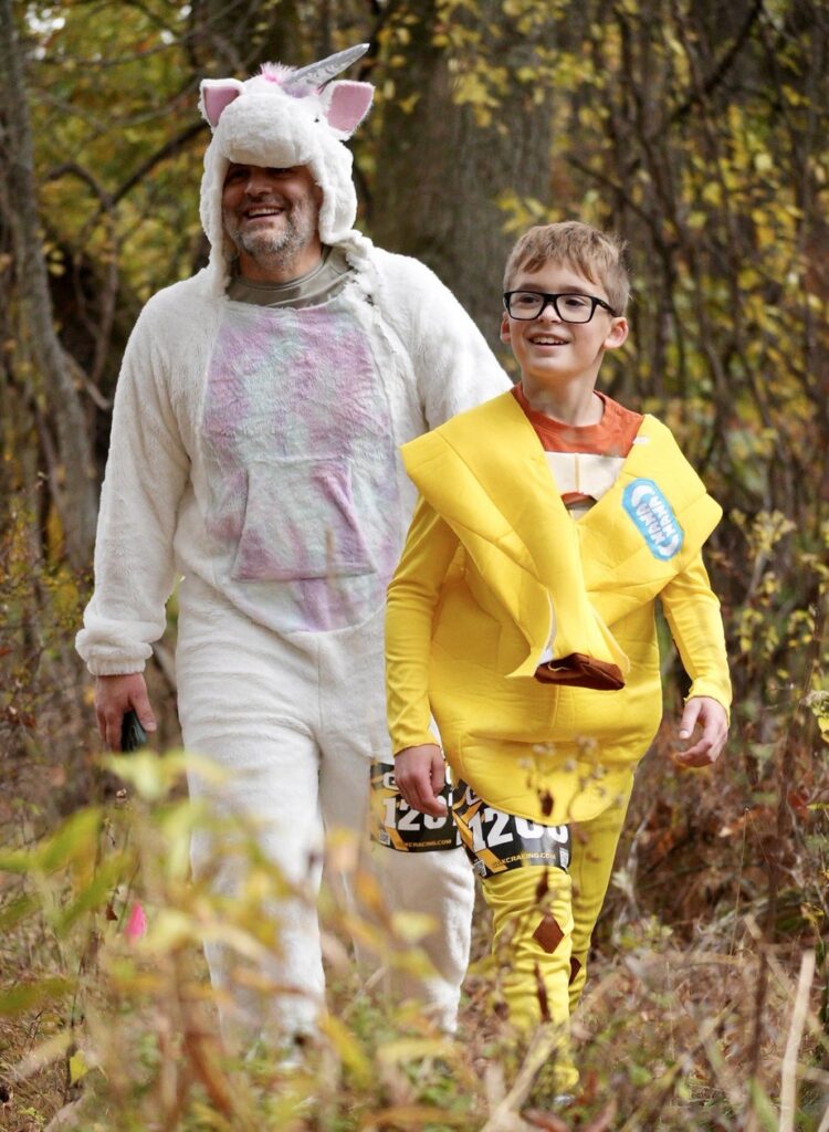 Man in unicorn costume and boy in banana costume walk in fall woods.