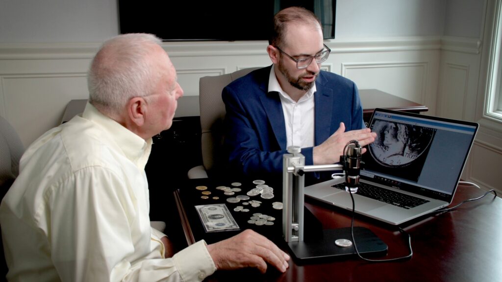 Two men examine coins on a laptop screen with a microscope.