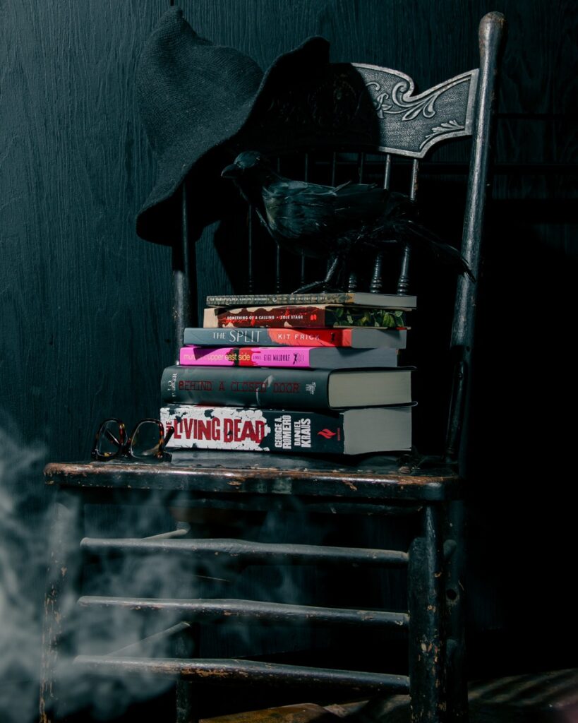 Stack of horror and thriller books by Pittsburgh authors on a chair with a crow.