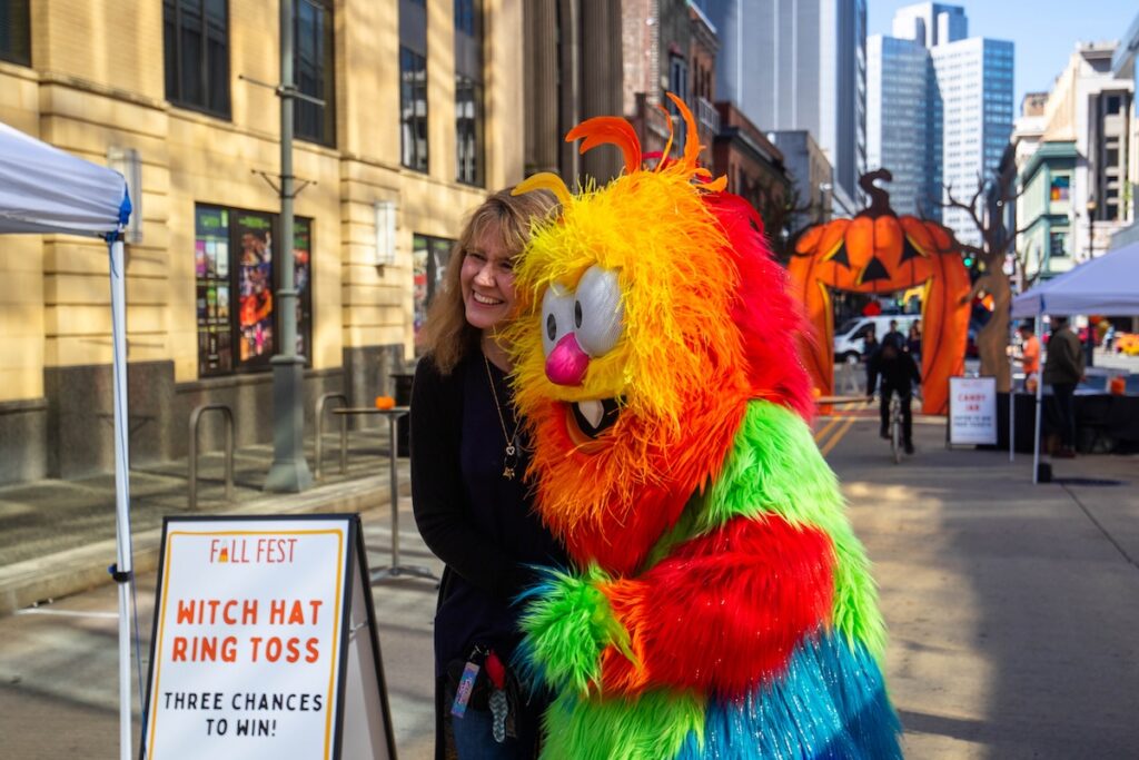 Woman poses with colorful monster mascot at Pittsburgh Playhouse Fall Fest witch hat ring toss.
