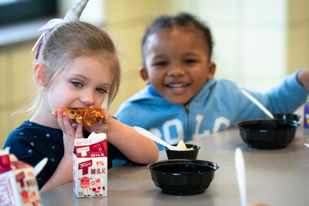 Young girl eats pizza, boy smiles, milk cartons on table