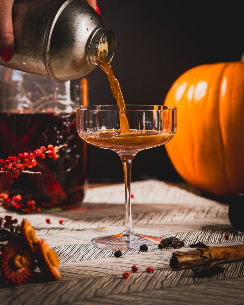 A person pours a drink from a cocktail shaker into a coupe glass in front of a jar of red liquid and a pumpkin.