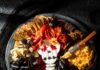 8 Spooky-Good Halloween Party Recipes for Home or Office Halloween cheese board with a brie skull, salami wig, marigolds, blackberry spiders, black grapes, cheeses, crackers, and a bowl of savory pumpkin dip.