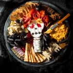 Halloween cheese board with a brie skull, salami wig, marigolds, blackberry spiders, black grapes, cheeses, crackers, and a bowl of savory pumpkin dip.
