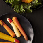 Carrot sticks on a black plate with red fingernails on them below two slices of cheese made to look like jack o lanterns.